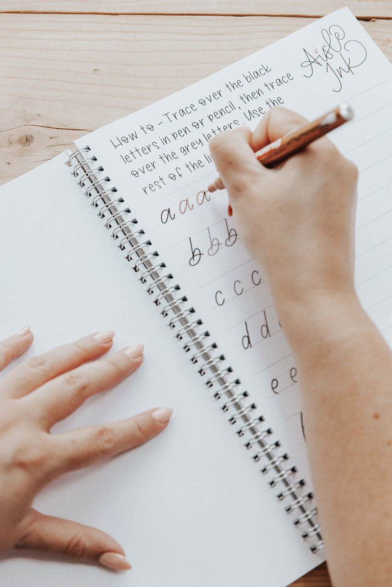 Person practicing handwriting the monoline font in the Aisle Ink workbook using a fine line rose gold pen