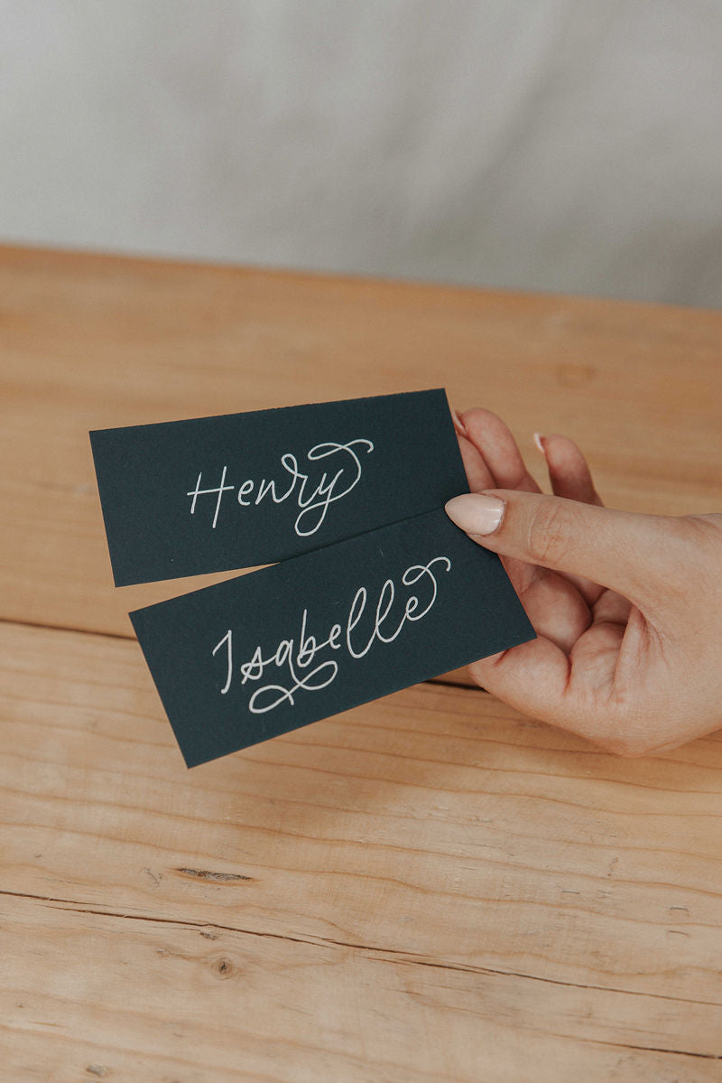 Hand holding two navy flat place cards using the monoline flourished font names on a wooden background