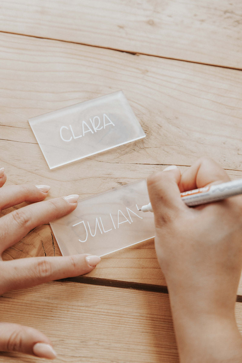 Hands writing on frosted acrylic name cards labelled 'Clara' and 'Julian' on a wooden surface.