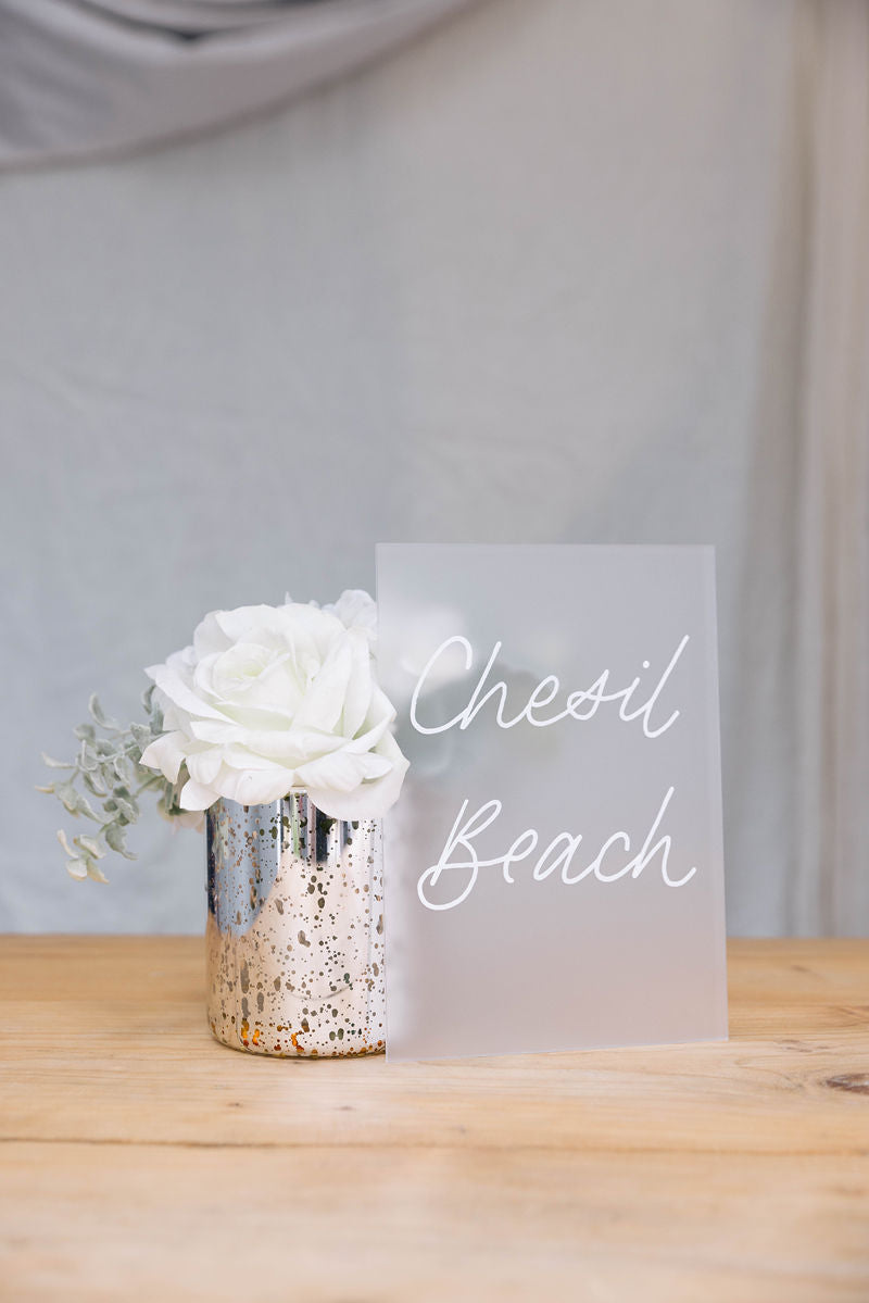 Floral arrangement in a silver vase with 'Chesil Beach' frosted acrylic Table Name on a wooden table.