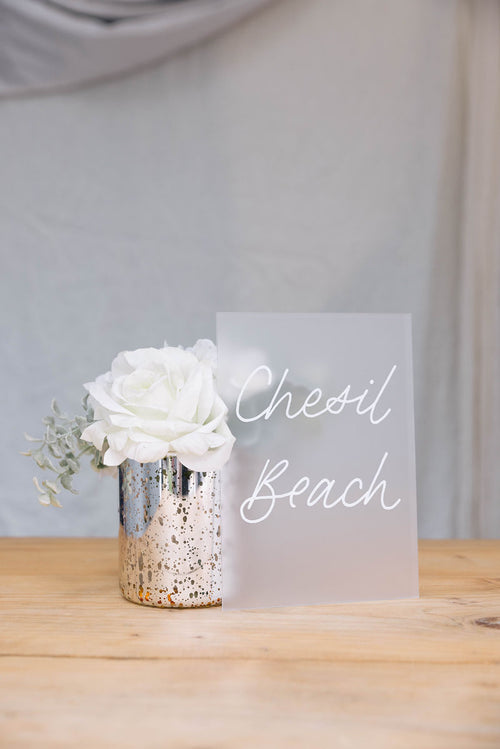 Floral arrangement in a silver vase with 'Chesil Beach' frosted acrylic Table Name on a wooden table.