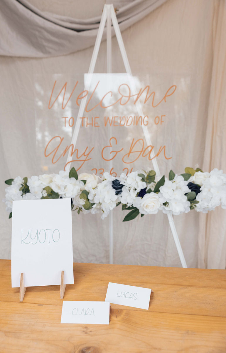 Clear acrylic welcome to the wedding sign with floral arrangement on a white easel, with white card place names written in green metallic ink and table name on a wooden table