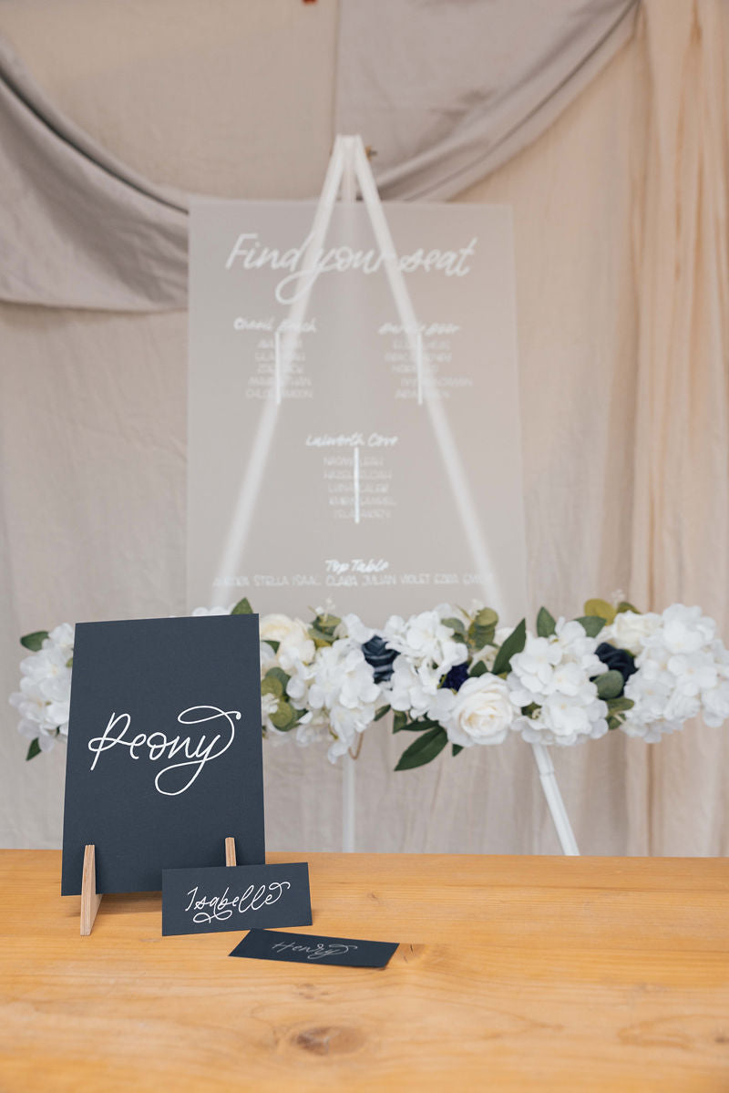 Frosted acrylic seating chart with floral arrangement on a white wooden easel, with navy blue place cards and table name in monoline flourished font